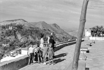 A group of children and two adults stand on a paved area with rustic benches, overlooking a scenic mountainous landscape. The scene has an old-fashioned, vintage feel with mountainous terrain in the background and small buildings scattered around. A leafless tree trunk is visible in the foreground.