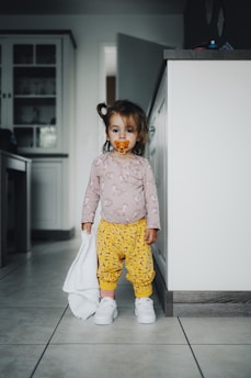 A toddler stands in a kitchen holding a small white towel. The child has a pacifier in their mouth and is dressed in a long-sleeve shirt with a unicorn print and yellow pants with small patterns, along with white sneakers. The background shows part of the kitchen with cabinets and countertops.