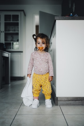 A toddler stands in a kitchen holding a small white towel. The child has a pacifier in their mouth and is dressed in a long-sleeve shirt with a unicorn print and yellow pants with small patterns, along with white sneakers. The background shows part of the kitchen with cabinets and countertops.