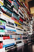 A large, towering collection of books stacked vertically in multiple piles, stretching into the distance on both sides. The books feature a variety of covers and thicknesses, representing diverse subjects and genres. The volume and arrangement suggest an overwhelming and impressive accumulation.