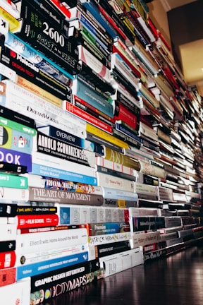 A large, towering collection of books stacked vertically in multiple piles, stretching into the distance on both sides. The books feature a variety of covers and thicknesses, representing diverse subjects and genres. The volume and arrangement suggest an overwhelming and impressive accumulation.