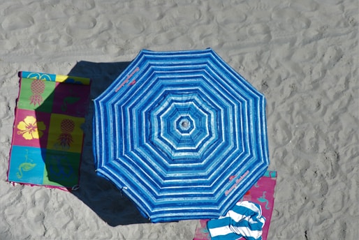 A vibrant blue striped beach umbrella is open on sandy ground, casting a shadow. Adjacent to it are colorful beach mats with floral and pineapple patterns, one partially covered by a blue and white striped towel.