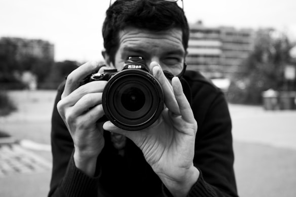 A person holds a Nikon camera, pointing it directly at the viewer, focusing on capturing an image. The background appears to be an urban setting with buildings and blurred surroundings, indicating shallow depth of field.