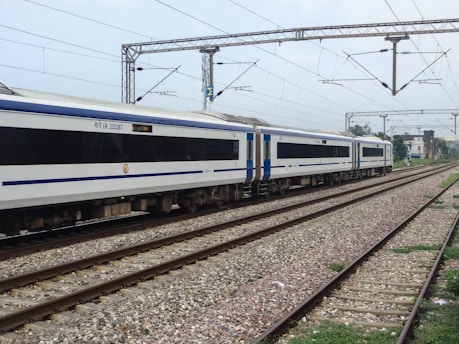 A modern train travels along a set of railway tracks. Overhead electric lines can be seen above the train, which is moving through a semi-urban area with greenery and a few buildings in the background. The train has a sleek design with a combination of white and blue colors.