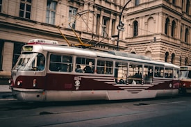A vintage-style red and white tram is traveling through an urban area with historic buildings. The tram is visible in profile with several passengers seated inside. Overhead power lines are visible, and the streets appear calm with no apparent traffic or pedestrians.