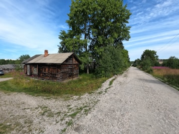 A rustic wooden house with a weathered roof stands beside a gravel road. The surroundings are lush with dense greenery, including a prominent large tree near the house. The sky is a bright blue with scattered white clouds. In the distance, more greenery lines the road, giving a sense of seclusion.
