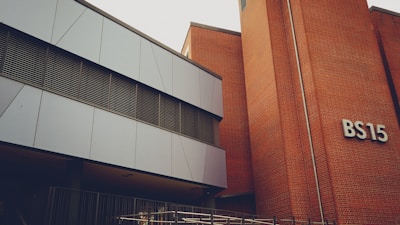A modern building with a mix of metal paneling and brickwork. The building has a sign labeled 'BS15' on the brick section. The metal panels are grey and the windows have horizontal metal frames. There's a railing at the lower part of the image, indicating a possible entrance or walkway.