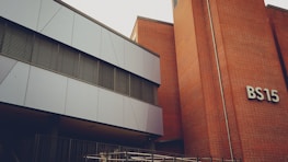 A modern building with a mix of metal paneling and brickwork. The building has a sign labeled 'BS15' on the brick section. The metal panels are grey and the windows have horizontal metal frames. There's a railing at the lower part of the image, indicating a possible entrance or walkway.