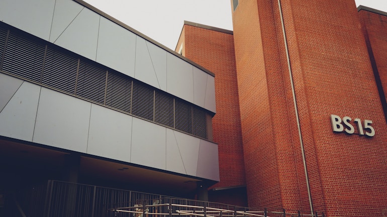 A modern building with a mix of metal paneling and brickwork. The building has a sign labeled 'BS15' on the brick section. The metal panels are grey and the windows have horizontal metal frames. There's a railing at the lower part of the image, indicating a possible entrance or walkway.