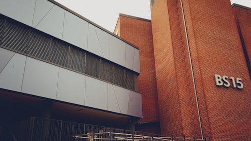 A modern building with a mix of metal paneling and brickwork. The building has a sign labeled 'BS15' on the brick section. The metal panels are grey and the windows have horizontal metal frames. There's a railing at the lower part of the image, indicating a possible entrance or walkway.