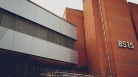 A modern building with a mix of metal paneling and brickwork. The building has a sign labeled 'BS15' on the brick section. The metal panels are grey and the windows have horizontal metal frames. There's a railing at the lower part of the image, indicating a possible entrance or walkway.