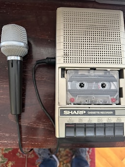 A vintage Sharp cassette recorder is placed on a wooden surface alongside a classic handheld microphone with a coiled cable. The cassette inserted in the recorder is visible through a transparent window. The buttons on the recorder include functions like record, play, and eject. Some patterned carpet is partially visible in the bottom of the image.