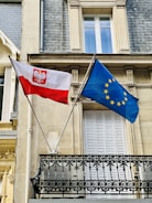 Two flags, one of Poland and the other of the European Union, are displayed on a building's facade. The flags are mounted on poles and positioned above an ornate black balcony railing with a beige stone wall and window in the background.