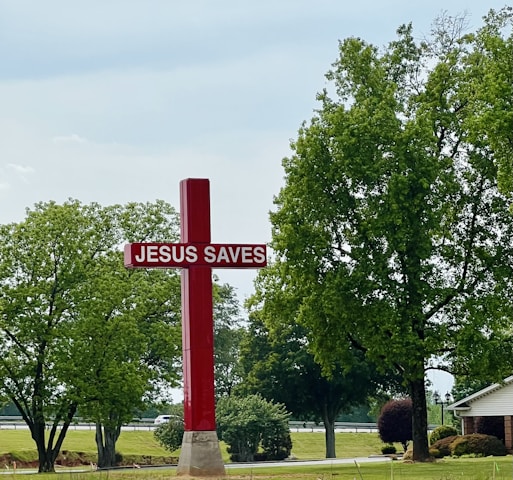 A large red cross with the text 'JESUS SAVES' stands prominently on a grassy area surrounded by trees. In the background, there is a road with a couple of vehicles visible. The sky is overcast, and a small house is partially visible to the right.