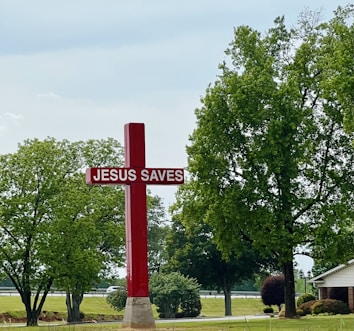 A large red cross with the text 'JESUS SAVES' stands prominently on a grassy area surrounded by trees. In the background, there is a road with a couple of vehicles visible. The sky is overcast, and a small house is partially visible to the right.