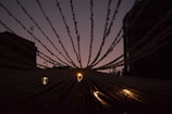 Strings of decorative lights stretch from a central point outward, illuminated against a dim sky. The scene is backlit, creating a silhouette effect with the buildings and banners. Three warm-toned street lamps provide soft light.