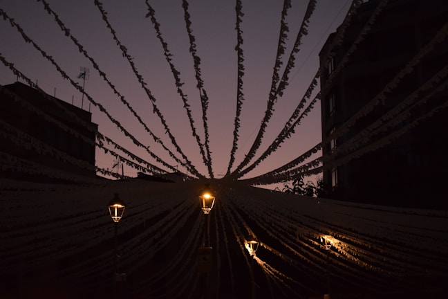 Strings of decorative lights stretch from a central point outward, illuminated against a dim sky. The scene is backlit, creating a silhouette effect with the buildings and banners. Three warm-toned street lamps provide soft light.