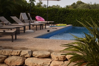 A tranquil swimming pool area with several lounge chairs arranged neatly alongside the pool. A pink inflatable ring and a couple of towels are on the chairs, suggesting recent use. The pool water is calm, and there's lush greenery surrounding the space, along with a sun umbrella providing shade.