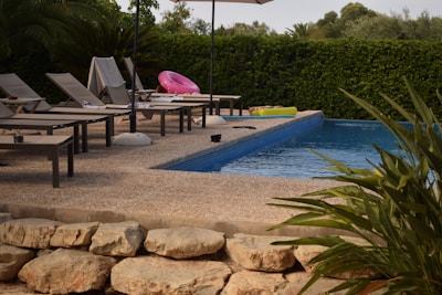 A tranquil swimming pool area with several lounge chairs arranged neatly alongside the pool. A pink inflatable ring and a couple of towels are on the chairs, suggesting recent use. The pool water is calm, and there's lush greenery surrounding the space, along with a sun umbrella providing shade.