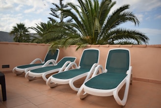 Four white and green lounge chairs are arranged on a tiled patio, surrounded by palm trees. The palm fronds create a tropical ambiance under a partly cloudy sky.
