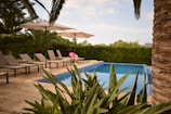 A serene outdoor setting with a small swimming pool surrounded by lush green plants and trees. Several beige lounge chairs are arranged neatly beside the pool under large white umbrellas. A pink inflatable float is visible on the pool's edge, adding a touch of color. The sky is partly cloudy, and the setting appears quiet and inviting.