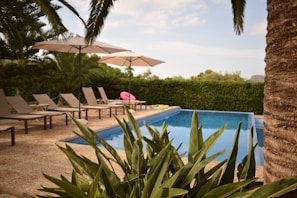 A serene outdoor setting with a small swimming pool surrounded by lush green plants and trees. Several beige lounge chairs are arranged neatly beside the pool under large white umbrellas. A pink inflatable float is visible on the pool's edge, adding a touch of color. The sky is partly cloudy, and the setting appears quiet and inviting.