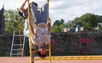 A person wearing a floral shirt is performing an acrobatic stunt, hanging upside down on a yellow ladder. The scene appears to be outside, with a backdrop of trees and a stone wall. Two red chairs are placed nearby on a paved surface under a partly cloudy sky.