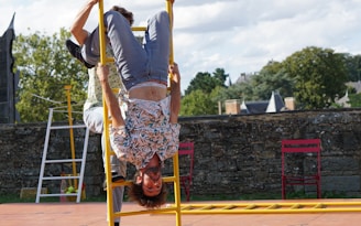 A person wearing a floral shirt is performing an acrobatic stunt, hanging upside down on a yellow ladder. The scene appears to be outside, with a backdrop of trees and a stone wall. Two red chairs are placed nearby on a paved surface under a partly cloudy sky.