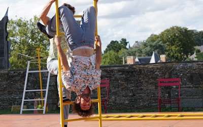 A person wearing a floral shirt is performing an acrobatic stunt, hanging upside down on a yellow ladder. The scene appears to be outside, with a backdrop of trees and a stone wall. Two red chairs are placed nearby on a paved surface under a partly cloudy sky.