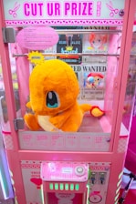 A bright pink arcade claw machine called 'Cut Ur Prize' features a large plush toy of a yellow and orange creature with blue eyes prominently displayed inside. The machine is adorned with colorful signage and instructions on how the game operates. Various 'Wanted' posters with cartoon characters are visible in the background.