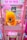 A bright pink arcade claw machine called 'Cut Ur Prize' features a large plush toy of a yellow and orange creature with blue eyes prominently displayed inside. The machine is adorned with colorful signage and instructions on how the game operates. Various 'Wanted' posters with cartoon characters are visible in the background.
