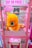 A bright pink arcade claw machine called 'Cut Ur Prize' features a large plush toy of a yellow and orange creature with blue eyes prominently displayed inside. The machine is adorned with colorful signage and instructions on how the game operates. Various 'Wanted' posters with cartoon characters are visible in the background.