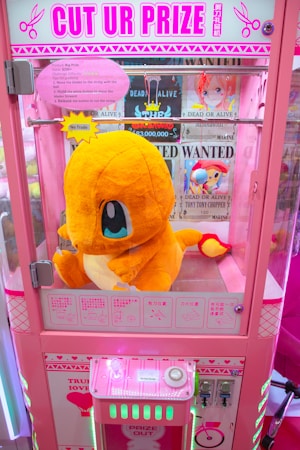 A bright pink arcade claw machine called 'Cut Ur Prize' features a large plush toy of a yellow and orange creature with blue eyes prominently displayed inside. The machine is adorned with colorful signage and instructions on how the game operates. Various 'Wanted' posters with cartoon characters are visible in the background.