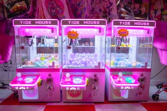 Three brightly colored claw machines lined up side by side, each with a neon pink hue. Inside, there are various prizes including plush toys and capsules with toys inside. The machines are set against a background of anime-themed merchandise.