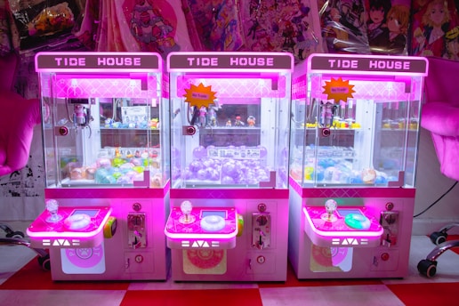 Three brightly colored claw machines lined up side by side, each with a neon pink hue. Inside, there are various prizes including plush toys and capsules with toys inside. The machines are set against a background of anime-themed merchandise.