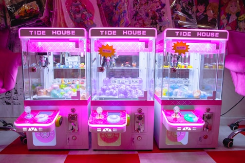 Three brightly colored claw machines lined up side by side, each with a neon pink hue. Inside, there are various prizes including plush toys and capsules with toys inside. The machines are set against a background of anime-themed merchandise.