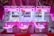 Three brightly colored claw machines lined up side by side, each with a neon pink hue. Inside, there are various prizes including plush toys and capsules with toys inside. The machines are set against a background of anime-themed merchandise.