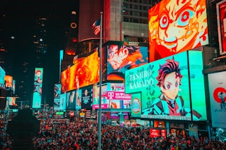 A bustling cityscape filled with large, colorful billboards displaying vibrant anime characters. The crowded street is packed with numerous people moving through the area. Bright neon lights dominate the environment, creating an energetic atmosphere. An American flag is partially visible atop one of the buildings.