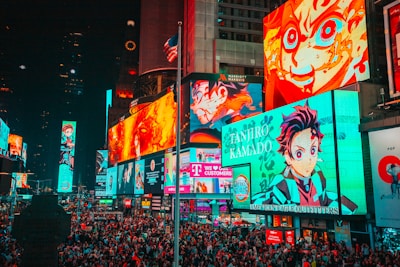 A bustling cityscape filled with large, colorful billboards displaying vibrant anime characters. The crowded street is packed with numerous people moving through the area. Bright neon lights dominate the environment, creating an energetic atmosphere. An American flag is partially visible atop one of the buildings.