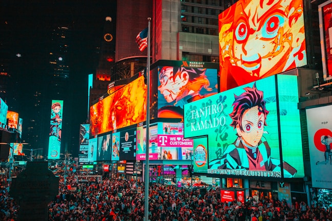 A bustling cityscape filled with large, colorful billboards displaying vibrant anime characters. The crowded street is packed with numerous people moving through the area. Bright neon lights dominate the environment, creating an energetic atmosphere. An American flag is partially visible atop one of the buildings.