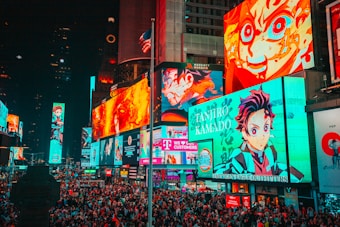 A bustling cityscape filled with large, colorful billboards displaying vibrant anime characters. The crowded street is packed with numerous people moving through the area. Bright neon lights dominate the environment, creating an energetic atmosphere. An American flag is partially visible atop one of the buildings.