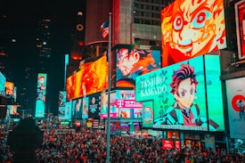 A bustling cityscape filled with large, colorful billboards displaying vibrant anime characters. The crowded street is packed with numerous people moving through the area. Bright neon lights dominate the environment, creating an energetic atmosphere. An American flag is partially visible atop one of the buildings.