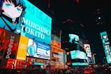 Brightly lit urban scene featuring animated characters on large digital billboards. The setting is a bustling city square with numerous advertisements from well-known brands such as McDonald's and Popeyes. Vibrant colors and large texts dominate the visual space, adding to the lively nightlife atmosphere. Cars and crowds fill the streets below, contributing to the dynamic energy.