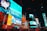 Brightly lit urban scene featuring animated characters on large digital billboards. The setting is a bustling city square with numerous advertisements from well-known brands such as McDonald's and Popeyes. Vibrant colors and large texts dominate the visual space, adding to the lively nightlife atmosphere. Cars and crowds fill the streets below, contributing to the dynamic energy.