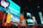 Brightly lit urban scene featuring animated characters on large digital billboards. The setting is a bustling city square with numerous advertisements from well-known brands such as McDonald's and Popeyes. Vibrant colors and large texts dominate the visual space, adding to the lively nightlife atmosphere. Cars and crowds fill the streets below, contributing to the dynamic energy.