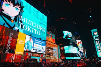 Brightly lit urban scene featuring animated characters on large digital billboards. The setting is a bustling city square with numerous advertisements from well-known brands such as McDonald's and Popeyes. Vibrant colors and large texts dominate the visual space, adding to the lively nightlife atmosphere. Cars and crowds fill the streets below, contributing to the dynamic energy.