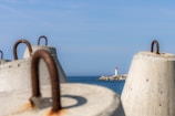 Large concrete barriers with rusty metal loops are positioned in the foreground, overlooking a calm sea. A small lighthouse with a red top is situated on a distant jetty amidst blue skies.
