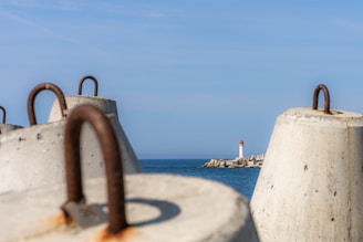 Large concrete barriers with rusty metal loops are positioned in the foreground, overlooking a calm sea. A small lighthouse with a red top is situated on a distant jetty amidst blue skies.