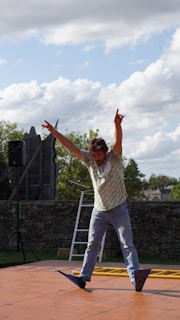 A person is performing a balancing act on a wooden platform outdoors. The individual stands with arms raised in a dynamic pose, surrounded by a partially cloudy sky. A ladder can be seen in the background along with a stone wall and some trees.