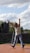 A person is performing a balancing act on a wooden platform outdoors. The individual stands with arms raised in a dynamic pose, surrounded by a partially cloudy sky. A ladder can be seen in the background along with a stone wall and some trees.